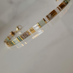 Puede incluir: Una pulsera delicada con un patr&oacute;n repetitivo de cuentas rectangulares en tonos blanco, verde p&aacute;lido y oro met&aacute;lico. El dise&ntilde;o incluye peque&ntilde;os espaciadores de tono dorado entre las cuentas y un cierre de mosquet&oacute;n dorado.