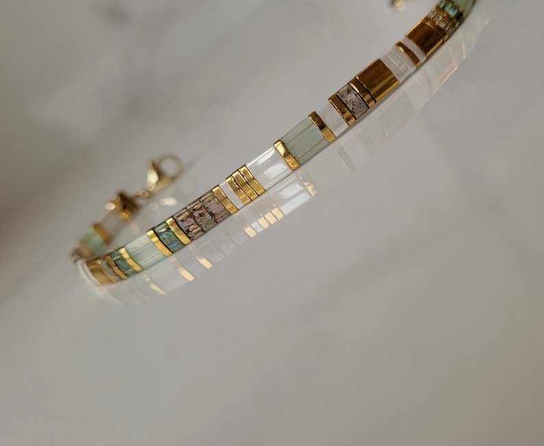 Puede incluir: Una pulsera delicada con un patr&oacute;n repetitivo de cuentas rectangulares en tonos blanco, verde p&aacute;lido y oro met&aacute;lico. El dise&ntilde;o incluye peque&ntilde;os espaciadores de tono dorado entre las cuentas y un cierre de mosquet&oacute;n dorado.