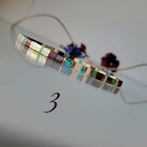 May include: A thin bracelet composed of small, rectangular, multi-colored beads in shades of turquoise, deep red, white, and metallic gold. The bracelet is positioned above the number 3, which is printed in a dark, elegant font.