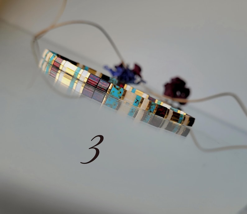 May include: A thin bracelet composed of small, rectangular, multi-colored beads in shades of turquoise, deep red, white, and metallic gold. The bracelet is positioned above the number 3, which is printed in a dark, elegant font.