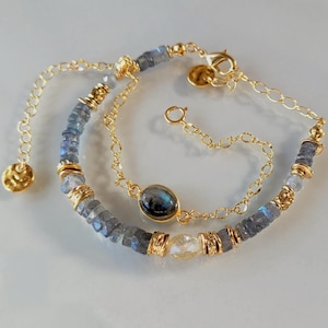 May include: A gold chain bracelet featuring faceted blue-grey labradorite beads, gold-toned spacers, and a central oval labradorite cabochon. The bracelet includes a delicate gold chain extension with a small, textured gold-toned circular charm.