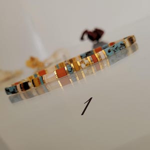 May include: A colorful bracelet with rectangular beads in shades of orange, turquoise, gold, and white. The bracelet is displayed on a reflective surface. The number "1" is written in black ink on the surface.