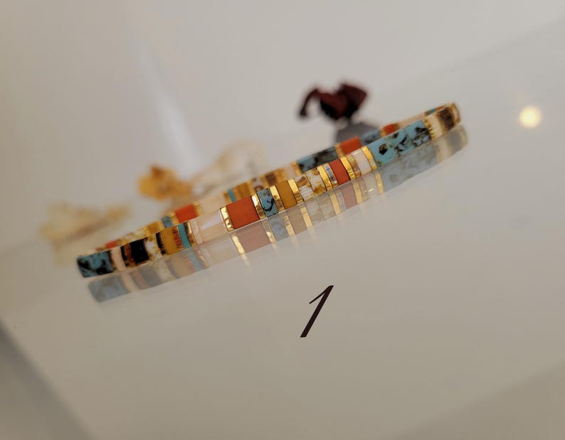 May include: A colorful bracelet with rectangular beads in shades of orange, turquoise, gold, and white. The bracelet is displayed on a reflective surface. The number "1" is written in black ink on the surface.