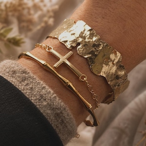 Peut inclure: Un ensemble de trois bracelets dorés. L'un est une manchette large avec une texture martelée irrégulière. Le deuxième est un bracelet à chaîne fine avec une croix centrale. Le troisième est un jonc rigide imitant une tige de bambou.