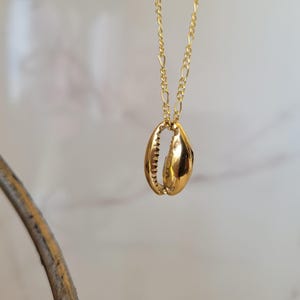 Op de afbeelding: Een goudkleurige ketting met een hanger in de vorm van een zeeschelp. De holle hanger heeft gedetailleerde randen en hangt aan een fijne ketting. De ketting wordt gepresenteerd tegen een zachte, neutrale achtergrond.