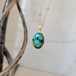May include: A gold-coloured necklace with a pendant featuring a turquoise stone. The oval-shaped stone has a faceted surface and is set in a gold-coloured bezel. The necklace chain is also gold-coloured.