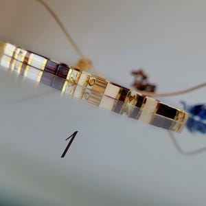 May include: A bracelet composed of rectangular beads in alternating colors of white, dark brown, and metallic gold. The bracelet features a thin, adjustable cord and is marked with the number 1 in a dark font on the surface below it.