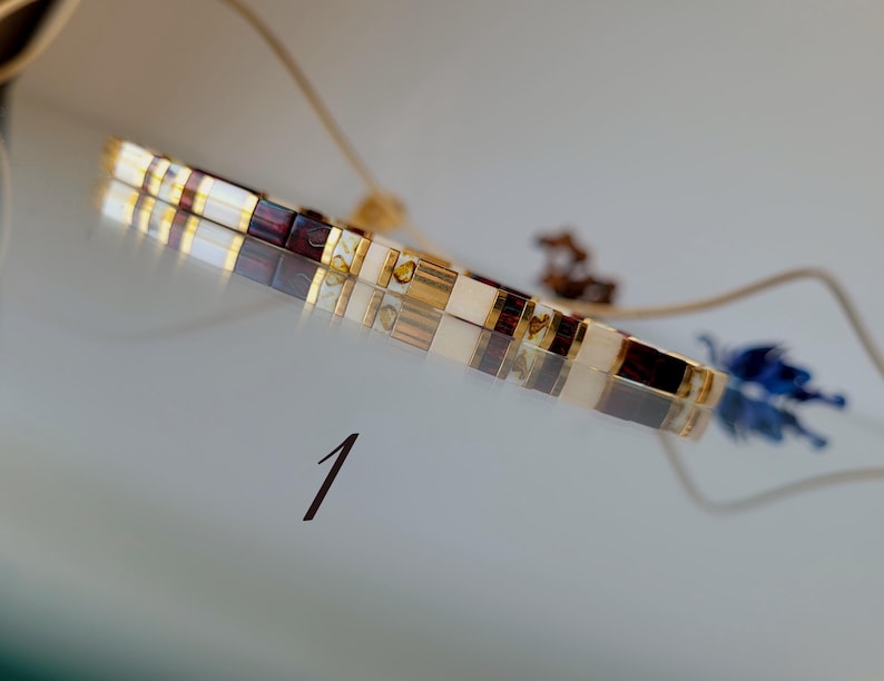 May include: A bracelet composed of rectangular beads in alternating colors of white, dark brown, and metallic gold. The bracelet features a thin, adjustable cord and is marked with the number 1 in a dark font on the surface below it.