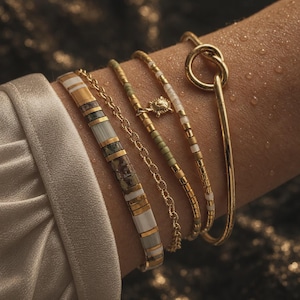 Peut inclure: Un ensemble de cinq bracelets dorés portés ensemble. La collection comprend un jonc lisse avec un détail de nœud, deux brins perlés avec de petits accents dorés, une fine chaîne dorée et un large bracelet composé de tuiles rectangulaires.