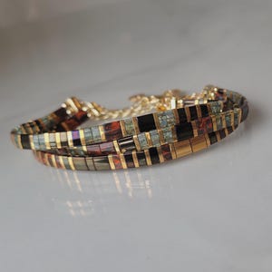 Può includere: Tre braccialetti impilati con un design a mosaico. I braccialetti presentano un mix di colori tra cui oro, nero, marrone e verde acqua. I braccialetti sono collegati da una catena d'oro.
