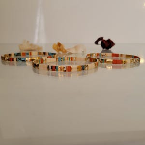 May include: Five beaded bracelets with a mix of colors including white, gold, turquoise, and red. The bracelets are displayed on a reflective surface, with dried flowers in the background. The bracelets are a delicate accessory.