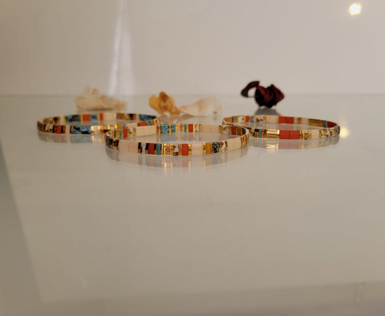 May include: Five beaded bracelets with a mix of colors including white, gold, turquoise, and red. The bracelets are displayed on a reflective surface, with dried flowers in the background. The bracelets are a delicate accessory.