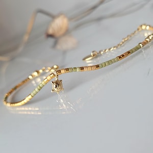Può includere: Un delicato braccialetto dorato con un piccolo ciondolo a forma di tartaruga. Il braccialetto presenta una catena e una sezione di perline con perline verde oliva e oro. Il ciondolo a forma di tartaruga è dorato e presenta dettagli intricati. Il braccialetto è su una superficie bianca.
