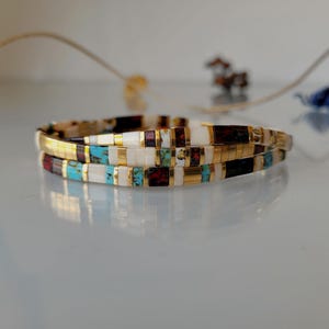 May include: A multi-layered bracelet composed of small rectangular beads in white, turquoise, dark red, and black, interspersed with thin gold-toned metallic spacers. The beads are arranged in a repeating pattern, creating a slender, flexible band.