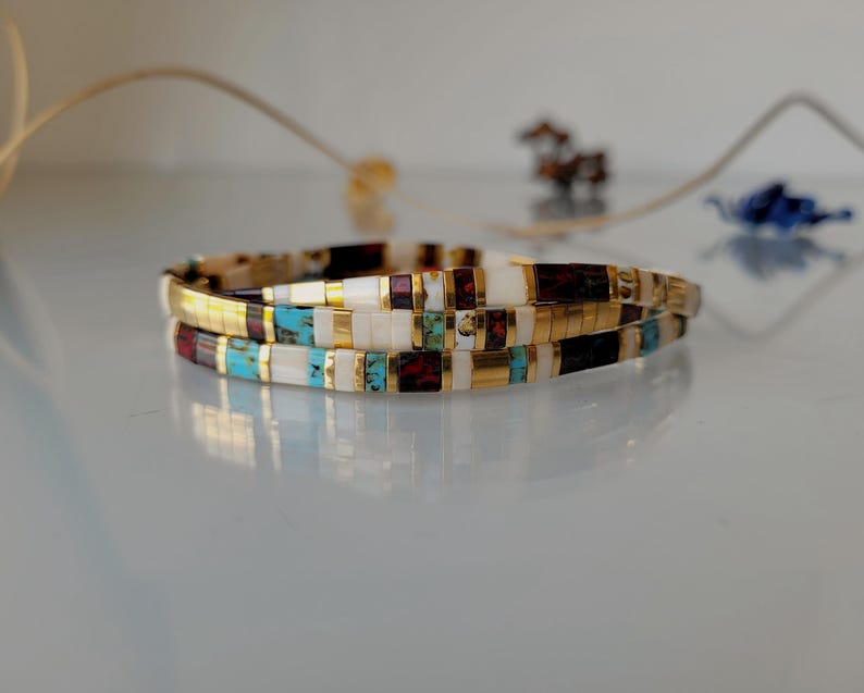 May include: A multi-layered bracelet composed of small rectangular beads in white, turquoise, dark red, and black, interspersed with thin gold-toned metallic spacers. The beads are arranged in a repeating pattern, creating a slender, flexible band.