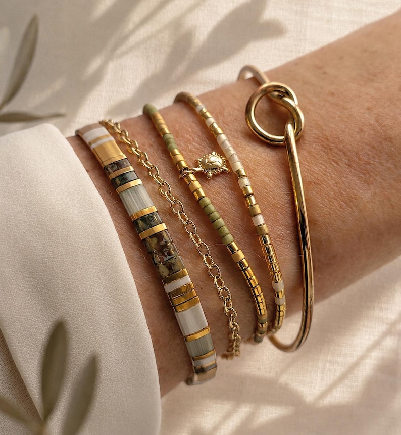 Puede incluir: Un conjunto de cinco pulseras que incluye una banda de cuentas planas con segmentos blancos, dorados y verde oscuro, una cadena dorada, un hilo de cuentas verdes y doradas con un peque&ntilde;o dije de tortuga, un hilo de cuentas blancas y doradas y un brazalete r&iacute;gido.