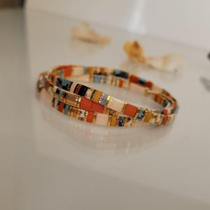 May include: A colorful beaded bracelet with a mix of orange, white, blue, and gold rectangular beads. The bracelet is made of multiple strands and is designed to wrap around the wrist. The beads are arranged in a repeating pattern.