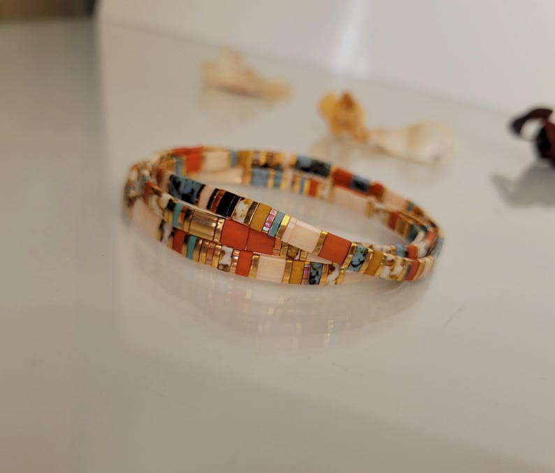 May include: A colorful beaded bracelet with a mix of orange, white, blue, and gold rectangular beads. The bracelet is made of multiple strands and is designed to wrap around the wrist. The beads are arranged in a repeating pattern.