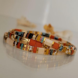 May include: Three colourful beaded bracelets. The bracelets feature a mix of rectangular beads in orange, cream, gold, black, turquoise, and yellow. The bracelets are stacked on top of each other, creating a layered effect.