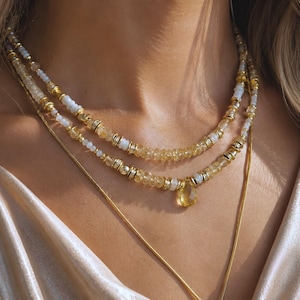 May include: A layered necklace set featuring two beaded strands and one thin gold-tone chain. The beaded strands consist of small, translucent yellow citrine-coloured stones, white beads, and gold-tone metal spacers, with a teardrop-shaped pendant.