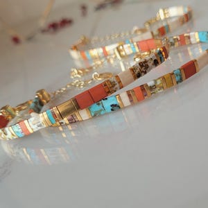 Könnte beinhalten: Zwei Armbänder mit Perlen, goldenen Verschlüssen und Ketten. Die Armbänder weisen ein Mosaikmuster aus rechteckigen Perlen in Rot-, Türkis-, Weiß- und Goldtönen auf. Sie werden auf einer reflektierenden Oberfläche präsentiert.
