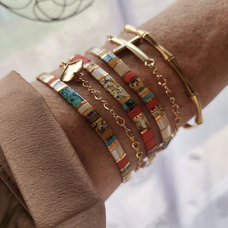 May include: A collection of gold bracelets and beaded bracelets. The bracelets include a gold bamboo-style bangle, a gold cross charm bracelet, and several beaded bracelets with colorful rectangular beads in shades of red, turquoise, and white.