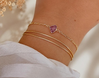 May include: A layered gold-tone bracelet set consisting of two thin snake chains and one delicate link chain featuring a single, faceted, triangular-shaped purple gemstone set in a gold-colored metal frame, worn around the wrist.