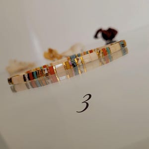 May include: A colorful bracelet with rectangular beads in shades of white, gold, red, blue, and yellow. The bracelet is lying on a reflective surface. The number "3" is visible below the bracelet.