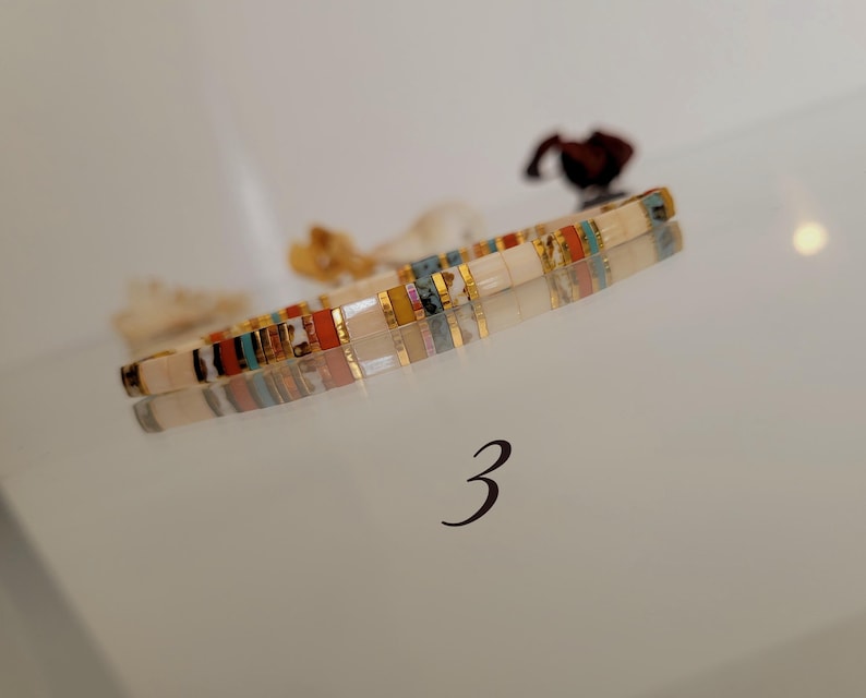 May include: A colorful bracelet with rectangular beads in shades of white, gold, red, blue, and yellow. The bracelet is lying on a reflective surface. The number "3" is visible below the bracelet.