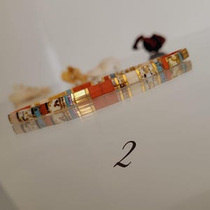 May include: A colorful bracelet with rectangular beads in shades of orange, white, gold, and blue. The bracelet is resting on a reflective surface, with the number "2" in the bottom right corner.