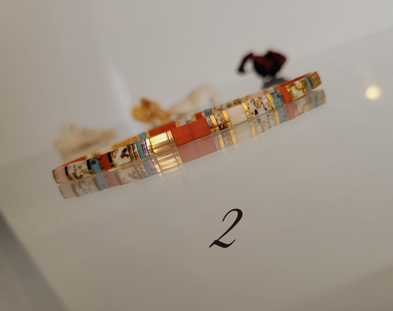 May include: A colorful bracelet with rectangular beads in shades of orange, white, gold, and blue. The bracelet is resting on a reflective surface, with the number "2" in the bottom right corner.