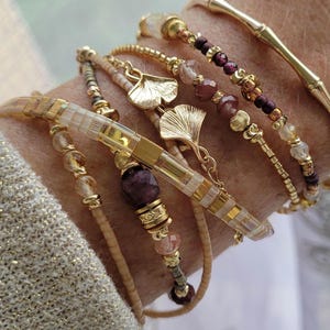 Puede incluir: Una colección de pulseras doradas con varios diseños. Algunas pulseras presentan cuentas en tonos marrón, morado y transparente. Una pulsera tiene un diseño similar al bambú, y otras incluyen dijes de hojas doradas. Las pulseras están apiladas.