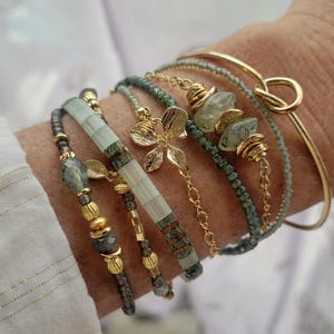 Può includere: Una collezione di braccialetti dorati e con perline in vari stili. I braccialetti presentano dettagli dorati, perline verdi e grigie e un ciondolo a forma di fiore dorato. Un braccialetto è un design a nodo dorato.