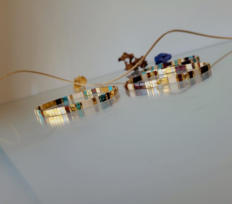 May include: Two thin bracelets made of small, square, multi-colored beads. The beads feature a mix of white, gold, turquoise, and dark brown tones arranged in a repeating pattern. The bracelets are circular and have a reflective, glossy finish.