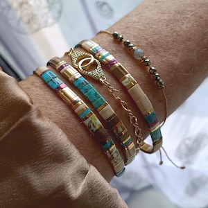 Puede incluir: Una colección de pulseras de cuentas en varios diseños. Las pulseras presentan cuentas turquesas, doradas y de tonos neutros. Una pulsera tiene un dije dorado. Las pulseras están apiladas en una muñeca, mostrando los diferentes estilos y colores.