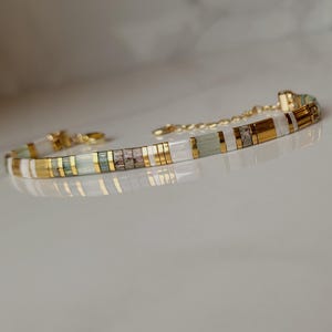 Puede incluir: Una pulsera delicada con un patr&oacute;n repetitivo de cuentas cil&iacute;ndricas en oro met&aacute;lico, verde p&aacute;lido transl&uacute;cido, blanco opaco y gris texturizado, terminada con una cadena extensora de metal dorado y un peque&ntilde;o cierre de mosquet&oacute;n.