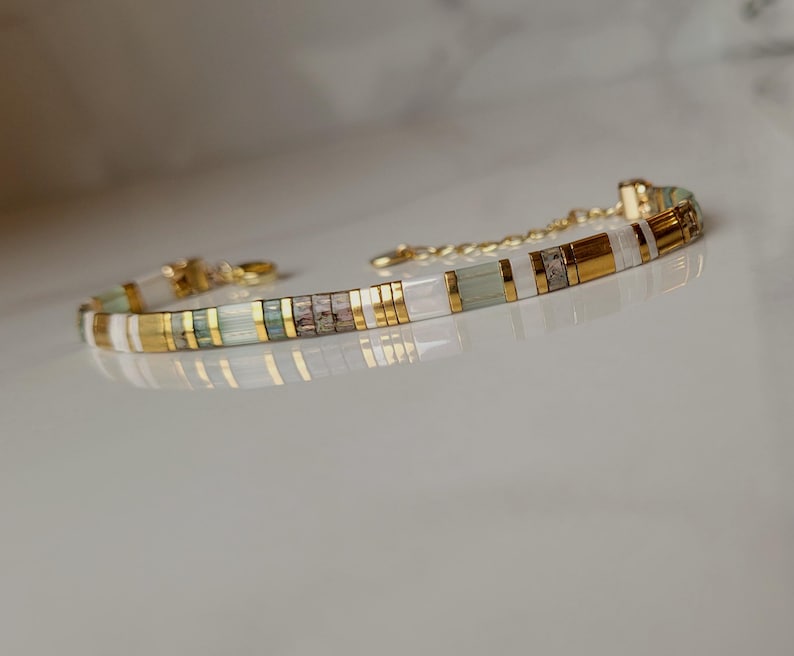 Puede incluir: Una pulsera delicada con un patr&oacute;n repetitivo de cuentas cil&iacute;ndricas en oro met&aacute;lico, verde p&aacute;lido transl&uacute;cido, blanco opaco y gris texturizado, terminada con una cadena extensora de metal dorado y un peque&ntilde;o cierre de mosquet&oacute;n.