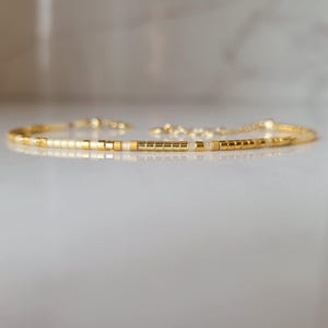 Op de afbeelding: Delicate goudkleurige armband met kleine, vierkante kralen in goud en gebroken wit. De armband heeft een verstelbare kettingsluiting. De kralen zijn geregen om een minimalistisch ontwerp te cre&euml;ren. Sieraden.