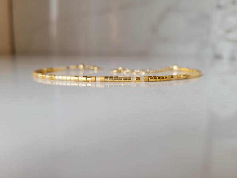 Op de afbeelding: Delicate goudkleurige armband met kleine, vierkante kralen in goud en gebroken wit. De armband heeft een verstelbare kettingsluiting. De kralen zijn geregen om een minimalistisch ontwerp te cre&euml;ren. Sieraden.