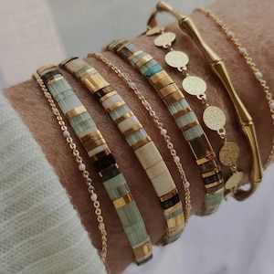 Puede incluir: Una colección de pulseras doradas y con cuentas en varios diseños. Las pulseras presentan detalles dorados, cuentas coloridas y cadenas delicadas. Las pulseras están apiladas en una muñeca, mostrando los diferentes estilos y texturas.