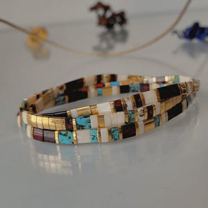 May include: A multi-strand bracelet composed of small rectangular beads in alternating colors of metallic gold, opaque white, turquoise blue, and dark maroon. The beads are arranged in a repeating pattern, creating a layered appearance.