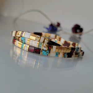 May include: A stack of two bracelets made of small rectangular beads in alternating colors of white, metallic gold, turquoise blue, and deep maroon. The beads are arranged in a repeating pattern, creating a multi-colored, segmented appearance.