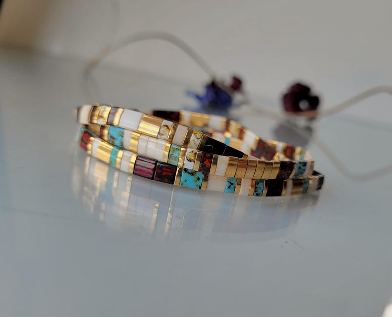 May include: A stack of two bracelets made of small rectangular beads in alternating colors of white, metallic gold, turquoise blue, and deep maroon. The beads are arranged in a repeating pattern, creating a multi-colored, segmented appearance.
