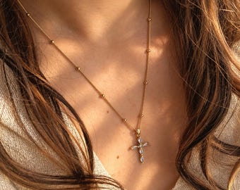 May include: A gold-toned chain necklace featuring small evenly spaced metal beads and a cross pendant. The cross is embellished with multiple clear, pear-shaped stones arranged in a symmetrical pattern, reflecting light against the gold metal.
