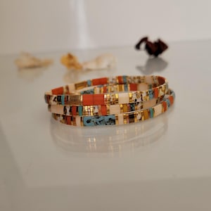 May include: Three colorful beaded bracelets stacked on top of each other. The bracelets feature a mix of orange, white, teal, and gold-colored beads. The bracelets are on a reflective surface.