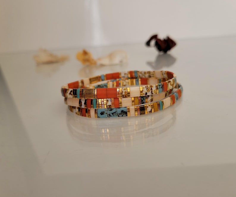 May include: Three colorful beaded bracelets stacked on top of each other. The bracelets feature a mix of orange, white, teal, and gold-colored beads. The bracelets are on a reflective surface.