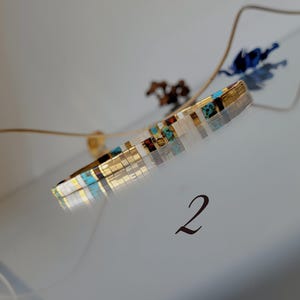 May include: A thin, cylindrical bracelet crafted from small, rectangular beads in a repeating pattern of metallic gold, white, turquoise, and dark brown. The bracelet is positioned at an angle, and the number 2 is printed below it.