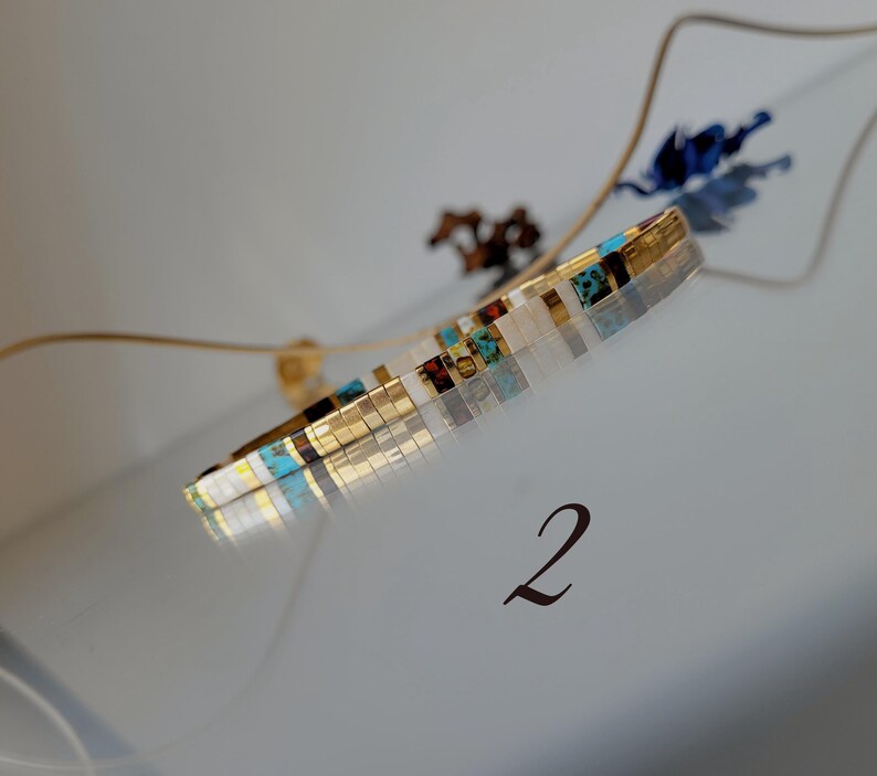 May include: A thin, cylindrical bracelet crafted from small, rectangular beads in a repeating pattern of metallic gold, white, turquoise, and dark brown. The bracelet is positioned at an angle, and the number 2 is printed below it.