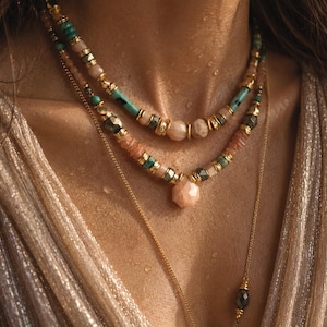May include: A layered necklace set featuring turquoise, peach-coloured, and metallic beads with gold-toned spacers. The design includes a central faceted peach-coloured stone pendant and a delicate gold-toned chain, creating a multi-strand appearance.