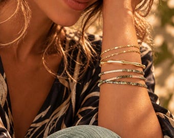 May include: A stack of five bracelets worn on the wrist. The set includes two thin gold chains, one gold bangle with a bamboo-like texture, and two beaded bracelets featuring small, multi-coloured green and cream-toned cylindrical beads.
